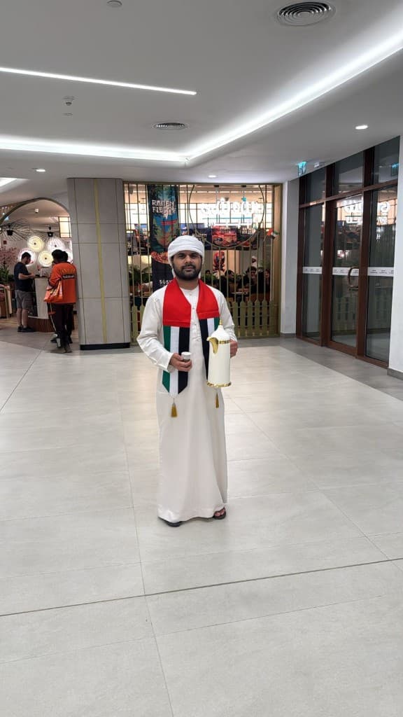 Host in kandura and UAE-flag scarf holding a white dallah and finjan in an Abu Dhabi mall