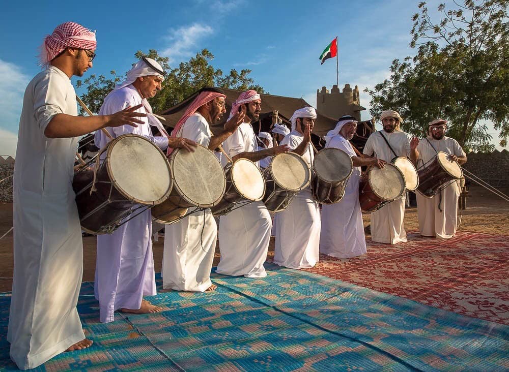 Men in kandura playing traditional drums on woven mats beside a heritage tent in Abu Dhabi