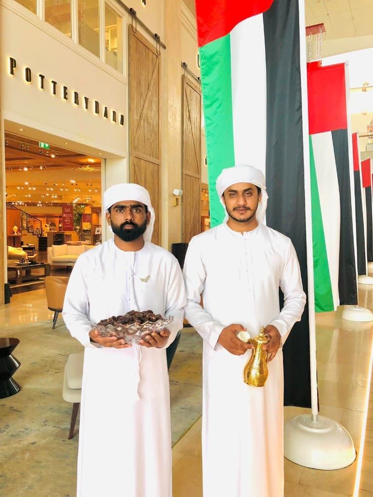 Emirati hospitality team serving dates and gahwa beside UAE flags in an Abu Dhabi shopping mall